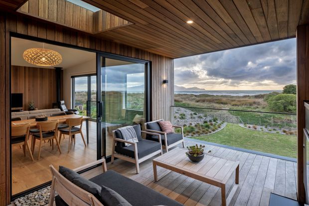 A contemporary indoor-outdoor living space with a timber-lined ceiling and walls, opening onto a covered timber deck through large glazed sliding doors. The foreground deck has lounge seating and a low timber coffee table, while the interior dining and living area is visible beyond. The home overlooks a coastal or lakeside landscape with water, dunes and distant hills.