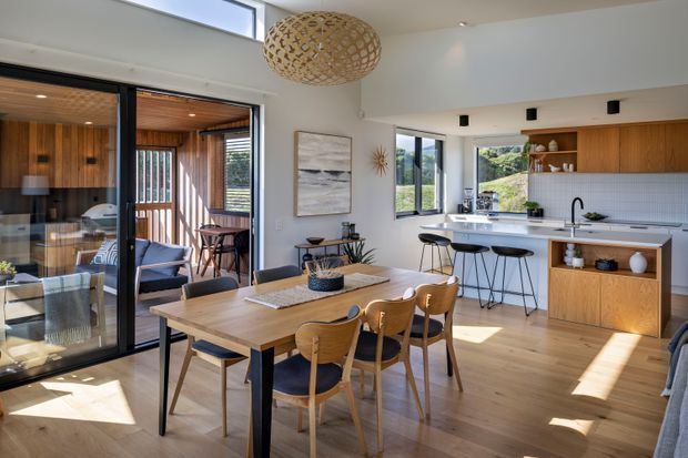 Bright open-plan dining and kitchen interior with warm timber flooring, a large timber dining table, and mid-century-inspired timber dining chairs. The kitchen features timber cabinetry, a pale benchtop, black-framed glazing, a breakfast bar with stools, and open shelving. A glazed opening connects the dining area to a timber-lined outdoor or secondary living space. A woven pendant light and contemporary artwork add texture and character.