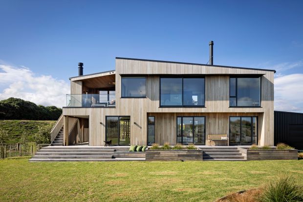 A contemporary two-storey house set in a rural or coastal landscape, photographed from the front across a broad lawn. The home features vertical timber cladding, large dark-framed windows, a low-pitched asymmetrical roof, multiple stepped timber decks and terraces, an upper-level balcony, and external stairs. A dark side volume and roof chimneys are visible, with expansive glazing providing views through the interior.