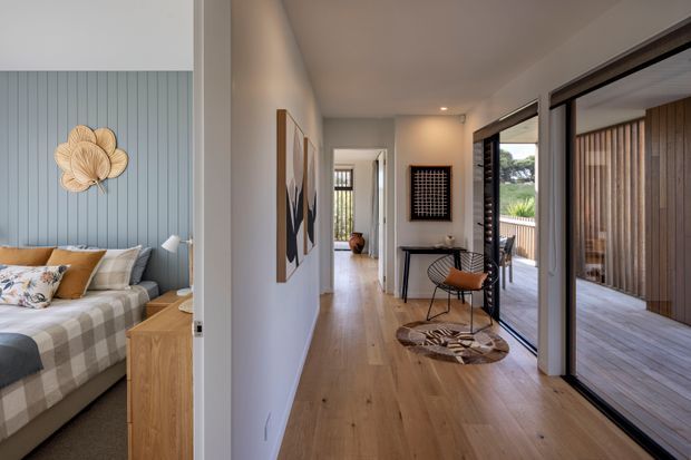 A bright contemporary residential hallway with warm timber flooring, white walls, and full-height glazed sliding doors opening to a covered timber-decked outdoor area. A small console, artwork, sculptural rug, and occasional chair furnish the corridor, while an adjoining bedroom is visible at left with a blue vertical-panel feature wall, upholstered bed, and timber bedside unit.