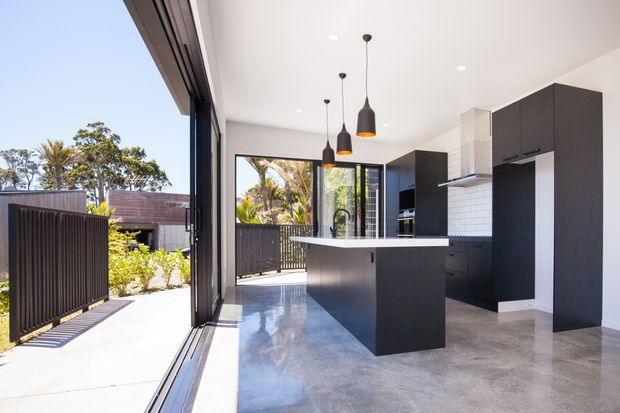 Bright contemporary kitchen with a large dark island, white stone-look benchtop, dark flat-panel cabinetry, white tiled splashback, pendant lighting, polished concrete flooring, and expansive sliding glass doors opening to a landscaped outdoor area.