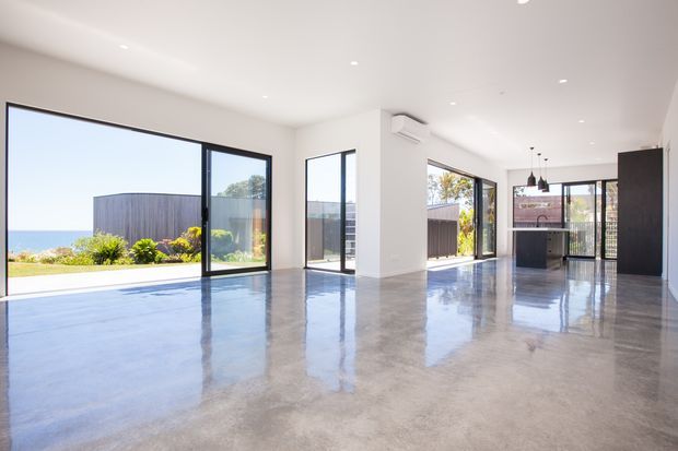 Bright, open-plan contemporary interior with polished concrete flooring, white walls and ceiling, extensive black-framed glazing, and sliding glass doors opening to a landscaped coastal outlook. A compact kitchen with a dark island and pendant lights is visible at the rear.