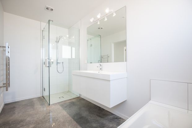 Bright contemporary bathroom with a frameless glass walk-in shower, wall-mounted white vanity with integrated basin, large mirrored cabinet, freestanding bathtub edge, and polished grey stone-look flooring. The room has white walls, recessed fittings, and a clean minimalist appearance.