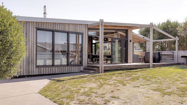 Single-storey contemporary home extension or pavilion viewed from the garden, featuring vertical grey timber cladding, large dark-framed glazed openings, and a covered timber outdoor terrace with exposed posts and beams. A paved path and lawn occupy the foreground, with built-in outdoor screening and a partially visible interior beyond the glazing.