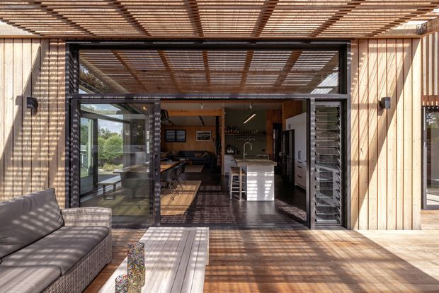 A bright contemporary timber-clad indoor-outdoor living space viewed from a covered deck or patio. Full-height black-framed glazed doors open into a combined living and kitchen area, with a central island, dark cabinetry, and lounge seating. A timber slatted pergola casts strong linear shadows across the deck and interior, while louvred side panels and extensive glazing reinforce the connection to the outdoors.