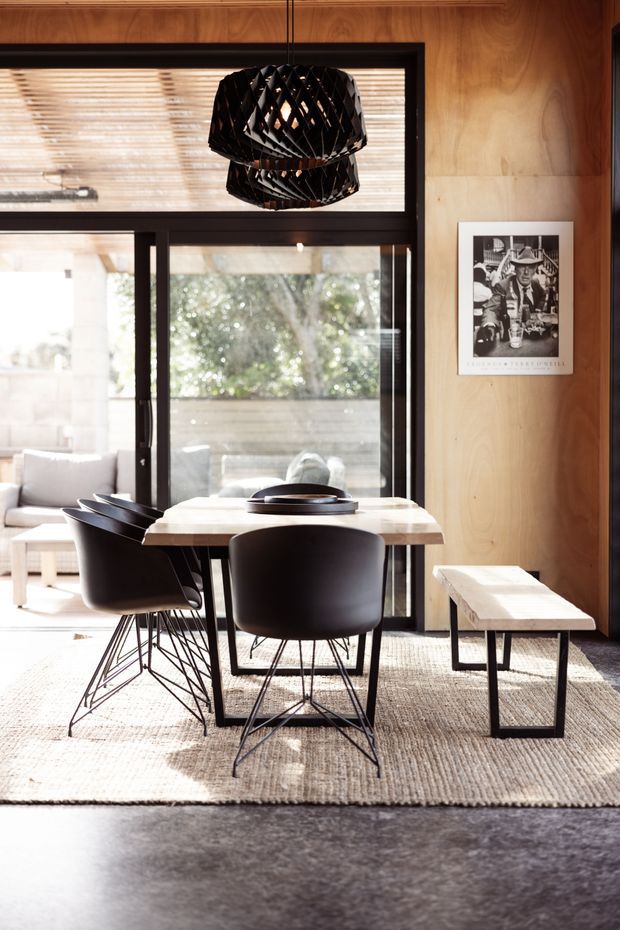 A warm, contemporary dining area with a light timber-lined wall and ceiling, large black-framed glazing, and a sculptural black pendant light. A pale rectangular dining table is surrounded by black molded chairs, with a matching light timber bench on a textured neutral rug. A bright living area and outdoor greenery are visible beyond the glazing, while framed artwork adds a small decorative accent.