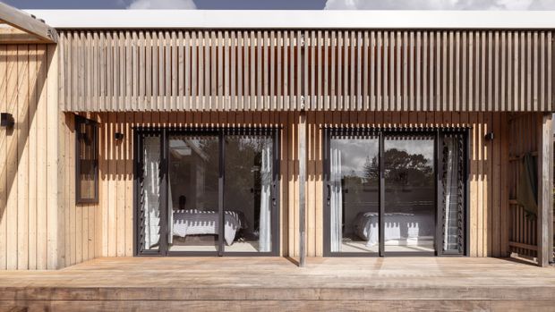 Exterior view of a contemporary timber-clad building with a broad timber deck, full-height glazed sliding doors, and a vertical timber slat screen providing shade and privacy. The glazing reveals a lightly furnished interior, while the image is primarily focused on the facade and outdoor deck.