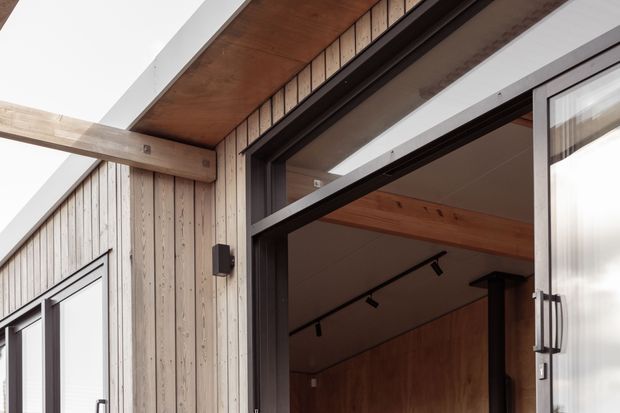 Close-up architectural view of a contemporary timber-clad building entrance, showing vertical weatherboard cladding, exposed timber framing and soffit, dark metal-framed glazing, and an open glazed doorway leading into a warm timber-lined interior with track lighting.