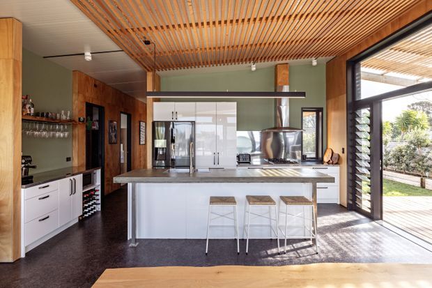 A spacious contemporary kitchen with a large white island and integrated seating, dark flooring, timber cabinetry, stainless-steel appliances, and a green feature wall. A timber slatted ceiling adds warmth and texture, while full-height glazed doors open to a timber deck and garden.