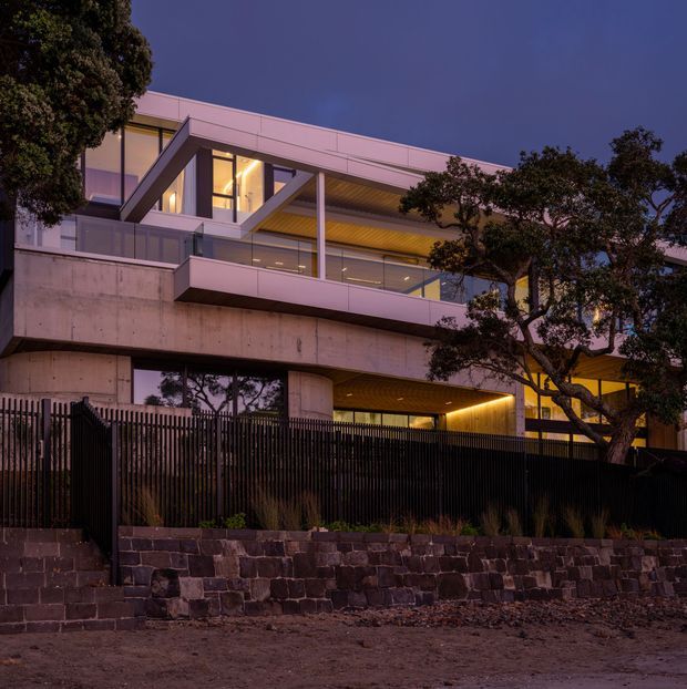 Twilight exterior view of a large contemporary multi-level residence with exposed concrete structure, extensive glazing, projecting upper-floor terraces, warm interior lighting, and a dark vertical-slat boundary fence above a stone retaining wall. Mature trees partially frame the building.
