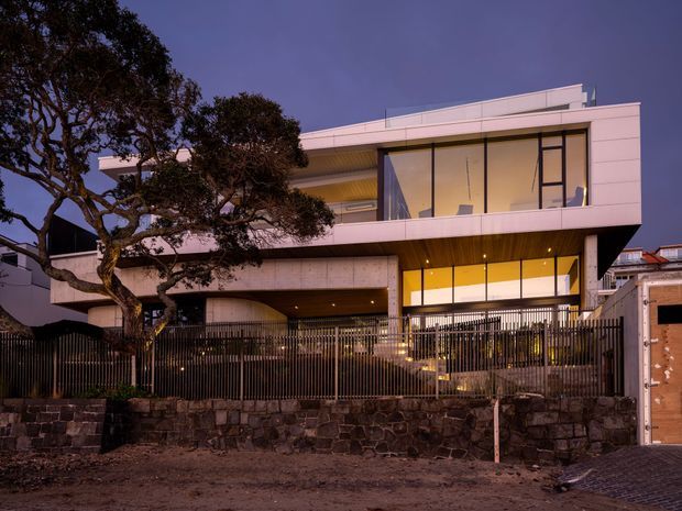 Twilight exterior view of a contemporary multi-level residence with strong horizontal lines, broad floor-to-ceiling glazing, projecting upper-floor volumes, a flat roof, and warm interior lighting. The building sits behind a dark metal fence and substantial stone retaining wall, with mature trees partially framing the facade.