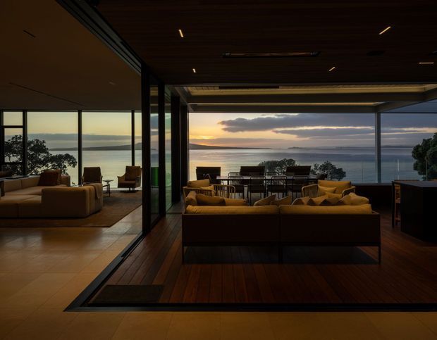 A sophisticated open-plan living and dining area with floor-to-ceiling glazing overlooking a calm coastal or waterfront landscape at sunset. The room features dark timber flooring and ceiling lining, warm neutral furnishings, low-profile sofas, a dining table, recessed linear lighting, and a seamless indoor-outdoor connection through expansive sliding glass walls. A secondary sitting room is visible through the glazed partition on the left.