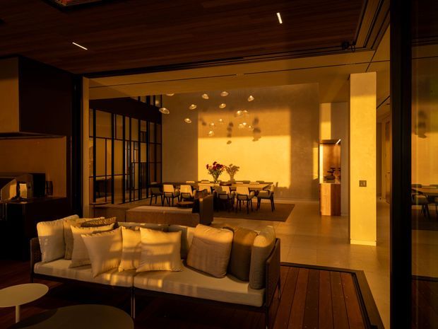 Warmly lit open-plan interior showing a living room in the foreground opening into a dining area. A large sectional sofa sits on a dark timber floor beside low round tables, while a dining table and chairs occupy the rear beneath sculptural pendant lights. Black-framed glazed partitions, a timber-lined ceiling, pale walls, and strong late-afternoon sunlight create a refined, intimate atmosphere.