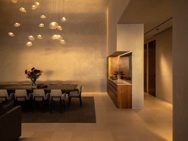 Warmly lit contemporary dining area with a long dark dining table and upholstered chairs, set against a textured beige feature wall. A sculptural cluster of pendant lights hangs above the table, while a timber built-in bar or buffet niche with integrated lighting sits beside a hallway. Large-format pale flooring and concealed architectural lighting create a refined, atmospheric interior.