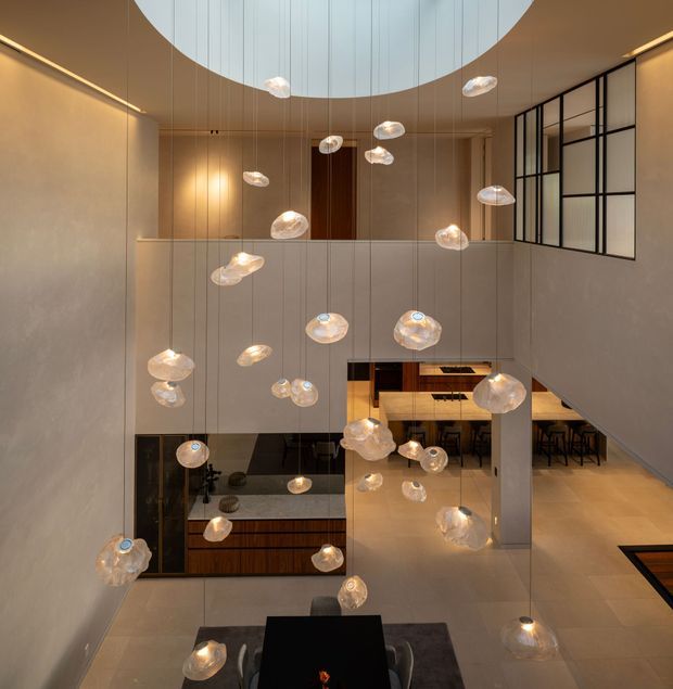 A dramatic double-height interior atrium with a large circular skylight, tall smooth plaster walls, and numerous suspended organic glass pendant lights cascading through the void. A dining table occupies the foreground, while a kitchen and additional seating areas are visible on the lower level and through the open-plan layout. Dark timber joinery and black-framed glazed panels contrast with the warm neutral finishes.