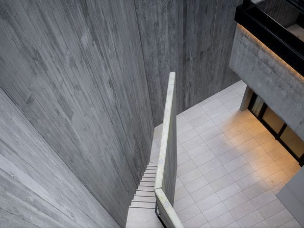 Looking down a narrow modern stairway beside tall grey textured concrete walls into a spacious tiled lower-level area. The stair has a simple light-coloured solid balustrade, while dark-framed glazing and a projecting concrete structure are visible across the void. The image has a cool, industrial architectural character with warm indirect lighting below.