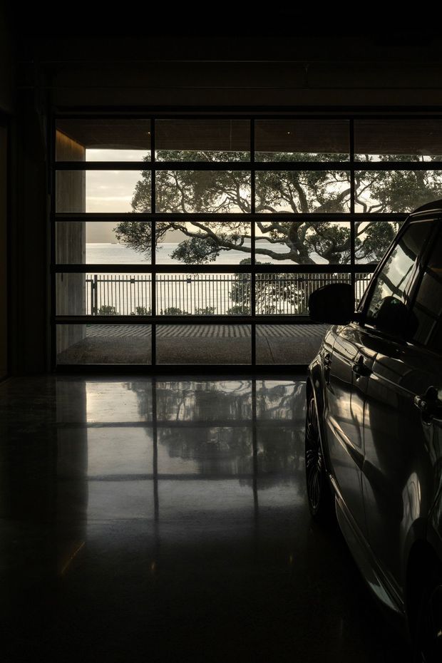 A dark, atmospheric garage interior photographed toward a large glazed sectional garage door. A car occupies the right side, while the reflective polished floor mirrors the strong rectangular window grid. Beyond the glazing are a balcony railing, mature tree, and a calm body of water, creating a dramatic indoor-outdoor connection.