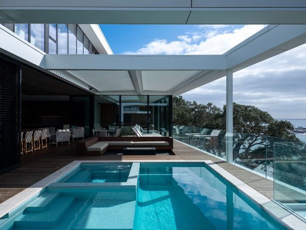A contemporary rooftop or elevated outdoor terrace with a swimming pool and integrated spa in the foreground. The space features expansive floor-to-ceiling glazing, a broad flat-roof canopy with slender white structural supports, dark exterior walls, timber decking, glass balustrades, and an outdoor lounge and dining area. Coastal water and trees are visible beyond the terrace.