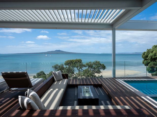 A contemporary elevated outdoor terrace overlooking a turquoise waterfront, with dark timber decking, built-in lounge seating, a low coffee table, glass balustrades, a small swimming pool, and a louvred pergola roof. The open structure frames a calm coastal view with strong horizontal lines and abundant daylight.
