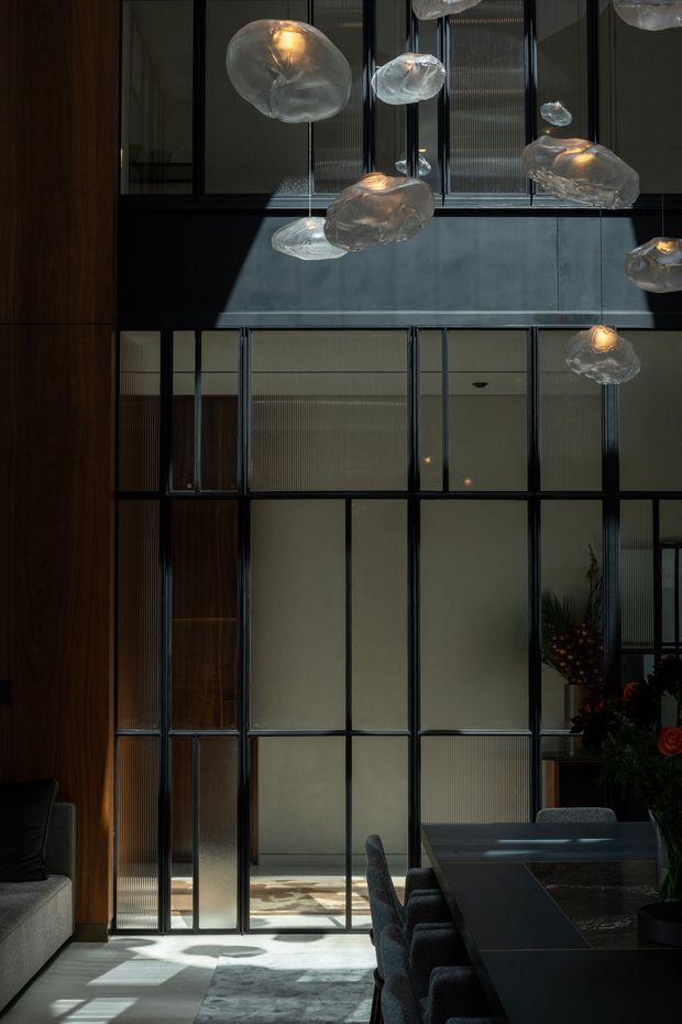 A dramatic double-height interior combining a dark dining area with a glazed, screened architectural partition. Tall black-framed translucent and ribbed glass panels divide the dining space from adjoining rooms, while sculptural translucent pendant lights hang overhead. Warm timber wall panelling, a dark dining table, upholstered chairs, and strong sunlight create a sophisticated, moody atmosphere.