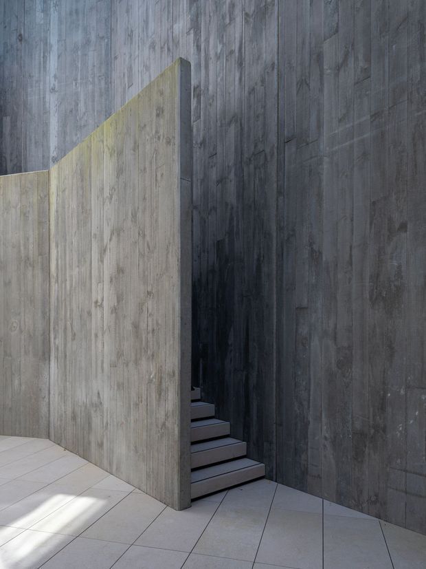 A minimalist architectural stairway enclosed by tall dark grey concrete walls. A lighter concrete partition forms a sculptural angled edge, concealing the stair flight. Large pale floor tiles contrast with the heavily textured vertical concrete surfaces, creating a stark, dramatic composition with strong natural light and shadow.