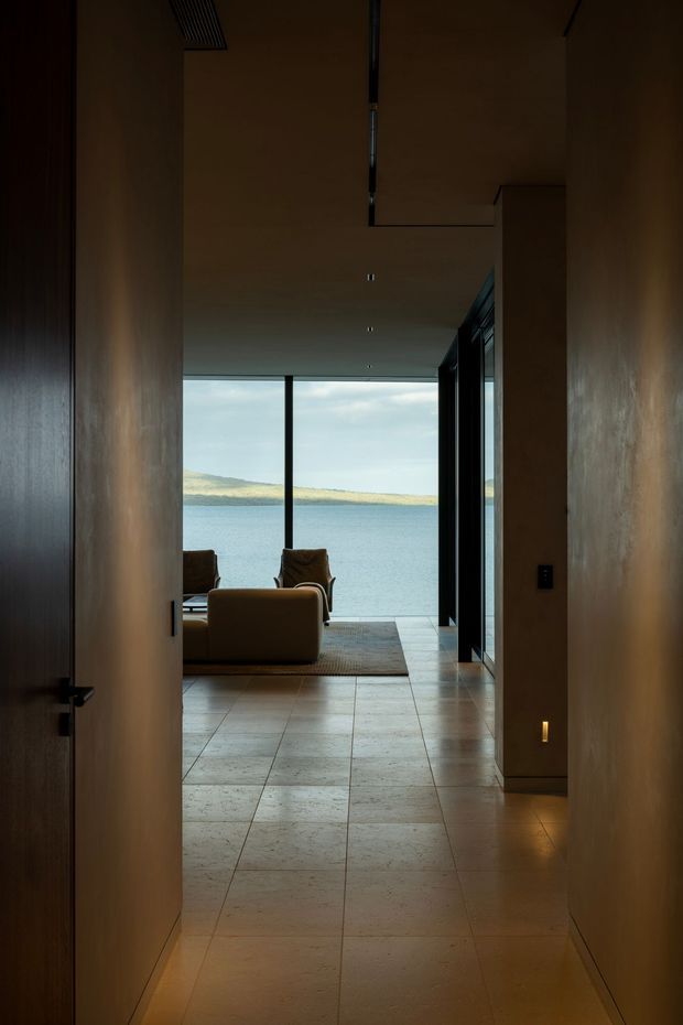 A moody interior view down a narrow hallway toward a waterfront living area. The corridor has warm dark timber wall or door panels, plastered walls, recessed ceiling lighting, and large pale stone or tile flooring. At the end, a low sofa and armchairs sit on a rug in front of full-height glazing with a calm body of water and distant shoreline beyond. A glazed or open doorway on the right leads toward the living space.