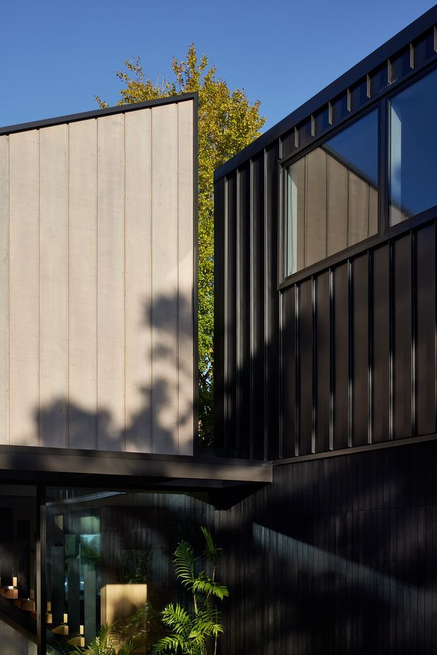 Exterior view of a contemporary multi-level building with strongly articulated black vertical cladding, dark metal framing, large glazed openings, and a contrasting pale concrete-clad upper volume. A covered entry or link passes beneath the projecting upper section, with planting visible near the entrance and a clear blue sky behind.