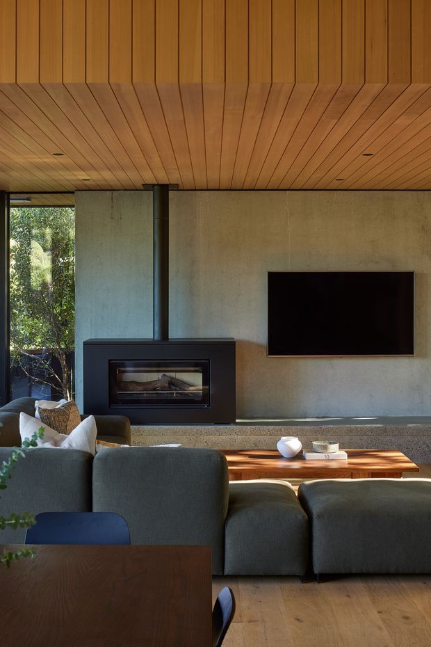 A warm, modern living room centered on a black built-in fireplace and wall-mounted television. The room features a richly timber-lined ceiling with angled plank detailing, a raw concrete feature wall and hearth, large windows overlooking greenery, dark upholstered sofas, and a low timber coffee table. The composition emphasizes natural materials, strong horizontal lines, and a connection to the surrounding landscape.
