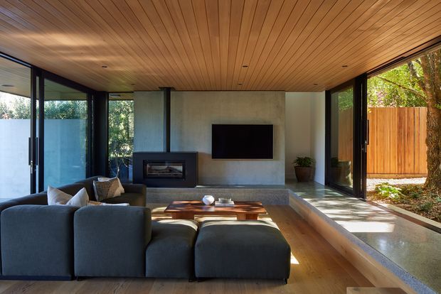 A contemporary living room with full-height glazed walls opening to a planted outdoor area. The room features a low dark sectional sofa, a timber coffee table, a built-in fireplace, a wall-mounted television, polished concrete surfaces, and a warm timber-lined ceiling. Black-framed sliding or folding glass doors create strong indoor-outdoor connection and abundant natural light.