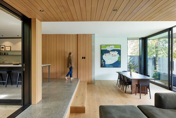 A refined open-plan dining and living area with a warm timber-lined ceiling, light timber flooring, polished concrete entry zone, and full-height black-framed glazing opening to an outdoor area. A dining table sits beside the glazing, while a contemporary lounge is visible in the foreground. A timber feature wall and concealed storage define the transition toward the kitchen.
