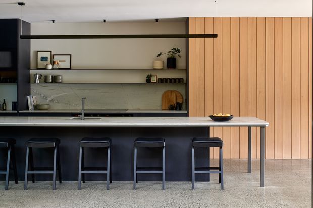 A contemporary open-plan kitchen dominated by a long dark island with five bar stools. The kitchen features matte dark cabinetry, a pale stone benchtop and splashback, open shelving, integrated appliances, black minimalist track lighting, and a full-height vertical timber-panelled wall. A terrazzo-like pale floor and restrained styling create a refined, warm modern interior.