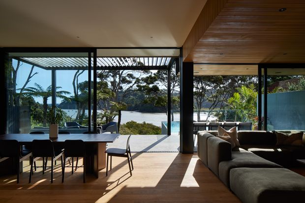 A light-filled open-plan living and dining area with full-height black-framed glazing opening toward a waterfront or poolside outlook. The room features warm timber flooring, a timber-lined ceiling with recessed lighting, a dark dining table and chairs, and a large neutral sectional sofa. External pergola slats cast strong linear shadows across the interior, creating a relaxed contemporary coastal atmosphere.