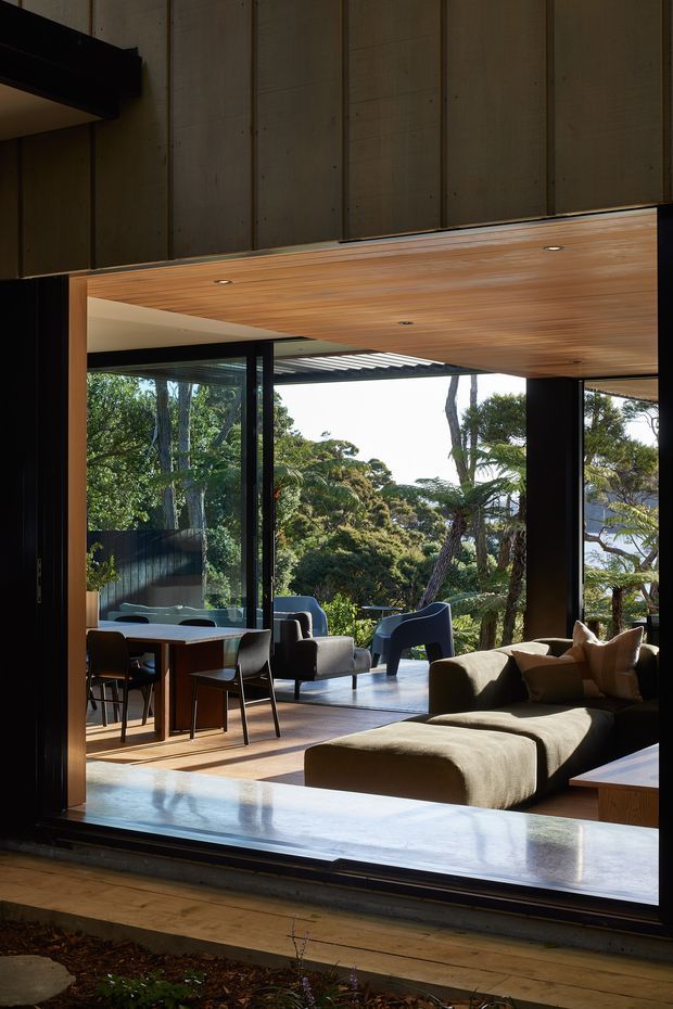 A refined open-plan living and dining area opening onto a leafy outdoor setting through extensive floor-to-ceiling glazing. The room features a large sectional sofa, dining table and chairs, dark structural framing, warm timber soffit, polished concrete or stone threshold, and strong indoor-outdoor connection. Natural light and mature vegetation create a calm, contemporary atmosphere.