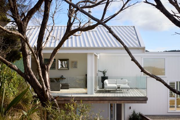 Exterior view of a light-coloured, contemporary multi-level home framed by mature tree branches. The building has white vertical cladding, a prominent pitched corrugated metal roof, large glazed openings, and an elevated timber deck furnished with a small outdoor seating arrangement. Garden planting and a lower-level doorway are visible.