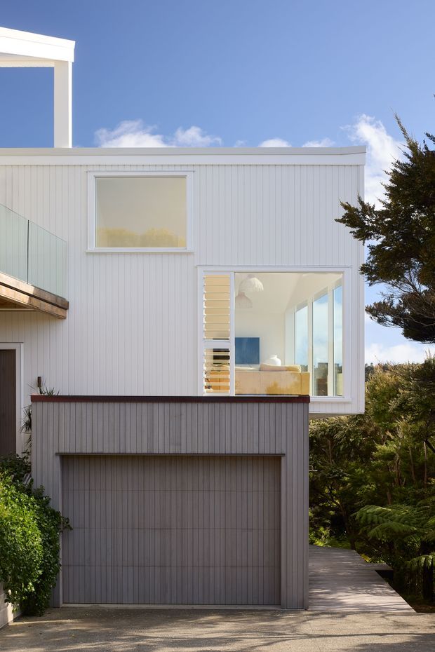 Exterior view of a compact contemporary multi-level house with white vertical cladding, a flat roofline, large glazed bedroom window, and an integrated garage at ground level. A timber walkway and dense planting frame the building, while a bright blue sky is visible above.