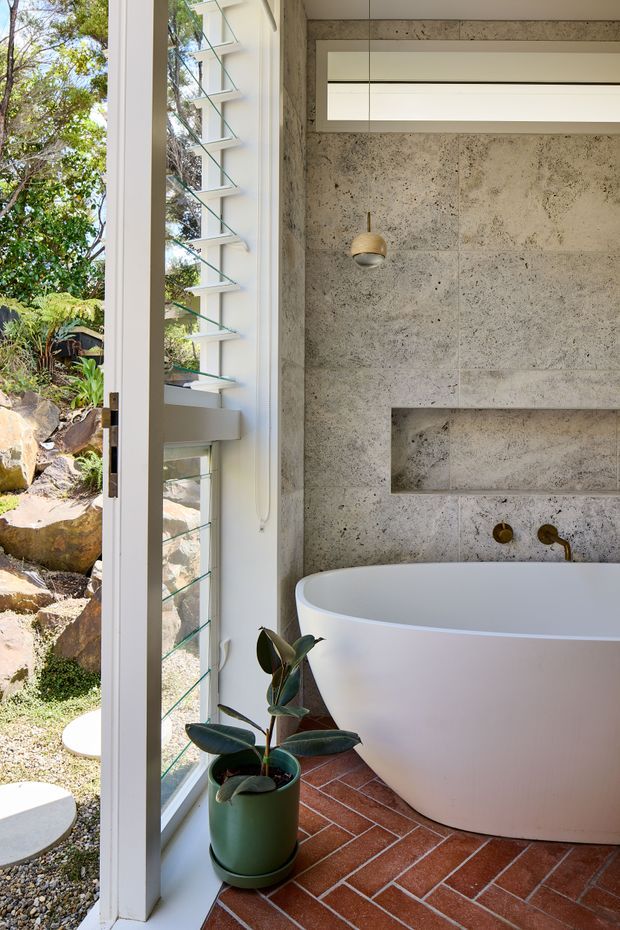 A bright, modern bathroom featuring a freestanding white bathtub set against a textured grey stone or concrete wall. The room has red terracotta brick flooring, a narrow recessed wall niche, a brass wall-mounted tap, a small pendant light, and a tall white-framed louvred window overlooking a landscaped rocky garden. A potted rubber plant adds greenery near the window.