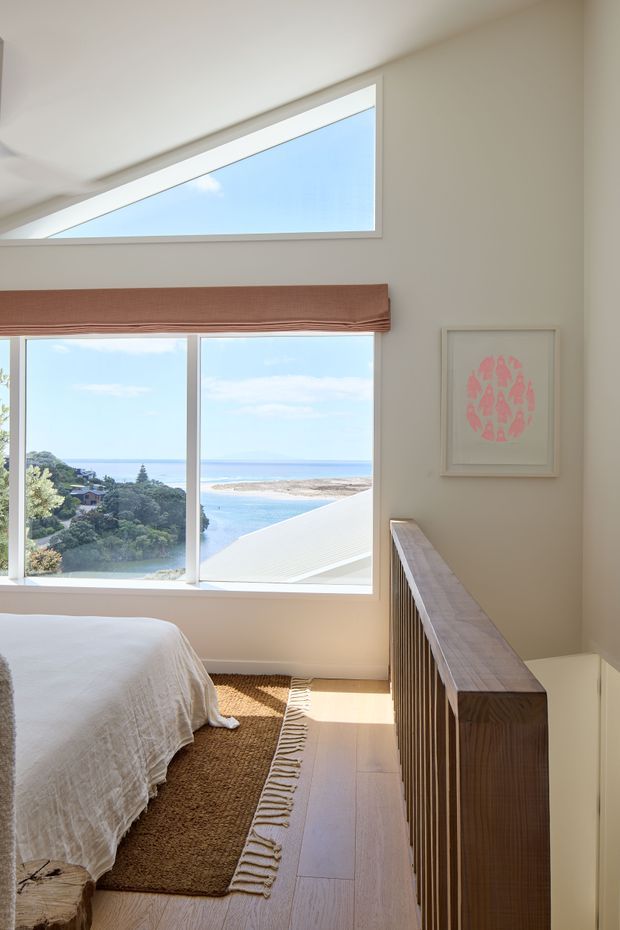 Bright coastal bedroom viewed from an upper-level landing or edge of the room, featuring a partially visible bed with white linen, a textured natural-fibre rug, pale timber flooring, a timber slatted balustrade, and large windows overlooking a bay or beach. A high triangular clerestory window brings additional daylight into the room. The palette is soft white, beige, pale timber, and muted terracotta-pink artwork.