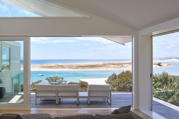 Bright contemporary living room opening onto a timber deck, with expansive floor-to-ceiling glazing framing a turquoise coastal inlet and sandy shoreline. Two outdoor lounge chairs sit on the deck, while the interior has a neutral palette, clean-lined furnishings, and a large skylight.