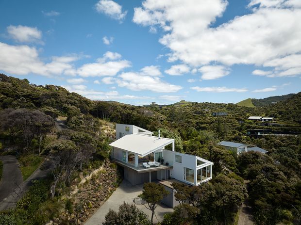 Aerial view of a modern multi-level home set within dense native bush on a hilly, sloped site. The residence has crisp white and pale grey geometric volumes, broad glazed openings, flat roof planes, and elevated terraces, with a driveway and retaining walls visible around the house. Other dispersed homes are visible among the surrounding vegetation.