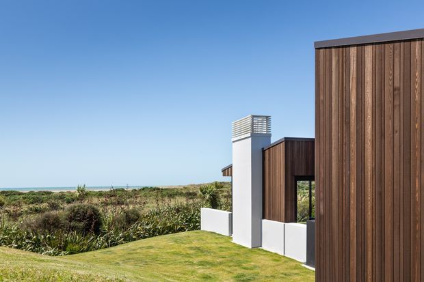 Exterior view of a contemporary coastal residence with strongly vertical dark timber cladding, crisp white rendered elements, and a modern chimney or ventilation tower capped with horizontal metal slats. The building sits beside low coastal vegetation and open ocean, with a manicured lawn in the foreground under a clear blue sky.