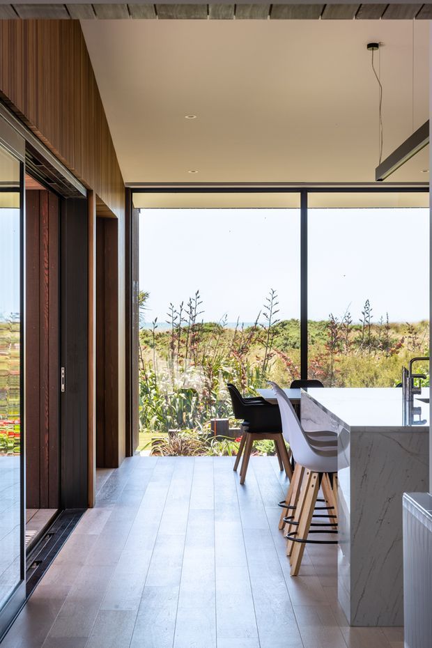 A bright, contemporary kitchen interior viewed toward full-height glazing and a landscaped coastal-looking garden. The room features a long marble-look island with integrated sink and bar seating, dark-framed glass doors and windows, warm vertical timber wall panelling, pale timber-look flooring, recessed ceiling lights, and a linear pendant light. The open glazing creates a strong indoor-outdoor connection.