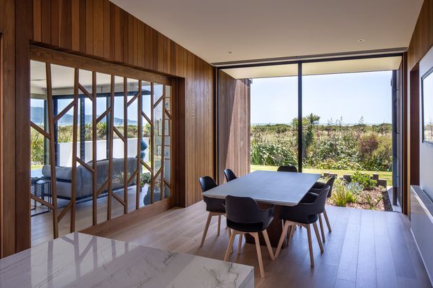 A contemporary dining area with a large rectangular dining table and dark upholstered chairs, opening to a landscaped garden through expansive floor-to-ceiling glazing. The room features rich vertical timber wall panelling, timber flooring, a pale marble-look kitchen island in the foreground, and a glazed internal partition with timber framing. Natural light and garden views create a warm, refined indoor-outdoor connection.