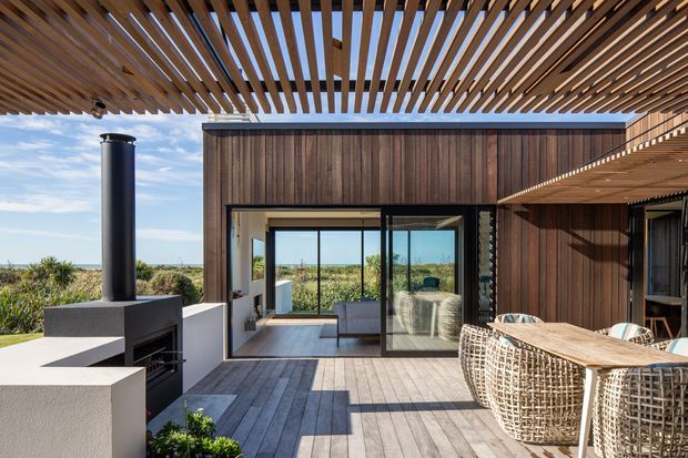 A contemporary outdoor entertaining area with a timber deck, slatted timber pergola, dark vertical timber-clad walls, and large sliding glass doors opening into the house. A built-in outdoor fireplace or chimney feature sits to the left, while a woven outdoor dining setting occupies the right side. The space overlooks low coastal vegetation and a distant ocean horizon.