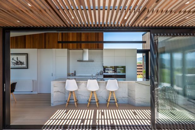 A bright contemporary kitchen viewed from an adjoining living area, featuring a large marble-look island with three white bar stools, white full-height cabinetry, a white subway-tile splashback, integrated appliances and timber wall panelling. A dramatic slatted timber pergola-style ceiling and matching overhead screen cast striped shadows across the timber floor, while large glazed sliding doors open to an outdoor view.