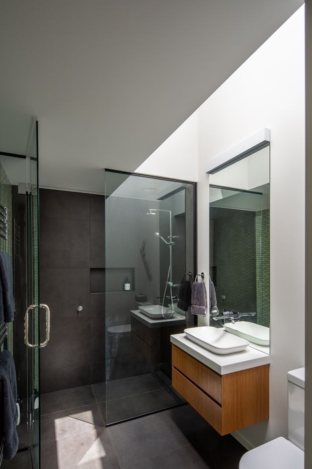 A contemporary bathroom with a walk-in glass shower enclosure, dark large-format wall and floor tiles, a floating timber vanity with a white countertop, twin vessel basins, a large mirrored wall, and integrated towel storage. The room has a clean, high-contrast palette with strong natural light entering from above or an adjacent opening.