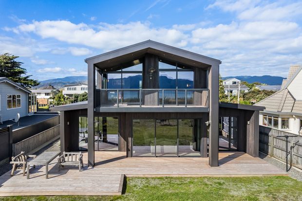 Rear exterior view of a contemporary two-storey residence with a prominent gabled roof form, dark vertical timber cladding, extensive glazing, and a broad timber deck. The upper balcony overlooks a landscaped backyard, with neighbouring homes and distant hills visible beyond.