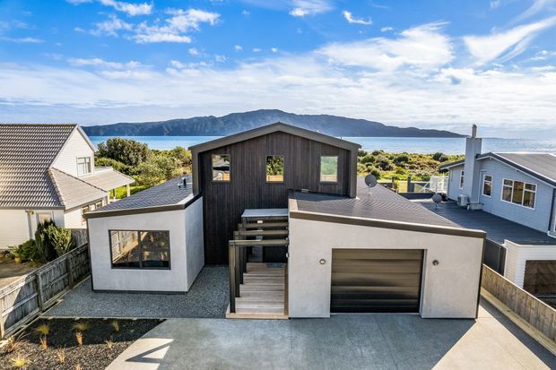 A contemporary multi-level coastal home viewed from above, with dark vertical timber cladding, white rendered walls, a flat-roofed garage volume, pitched roof elements, and a central outdoor stair and deck. The surrounding suburban homes, ocean inlet, and mountainous headland are visible in the background.