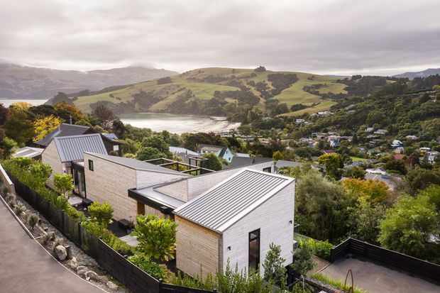 Elevated exterior view of a contemporary multi-volume house with pale horizontal cladding and several pitched metal roofs, set on a steep landscaped hillside overlooking a sheltered inlet and rolling green hills. The surrounding suburban settlement and dense planting reinforce the sloped coastal setting.