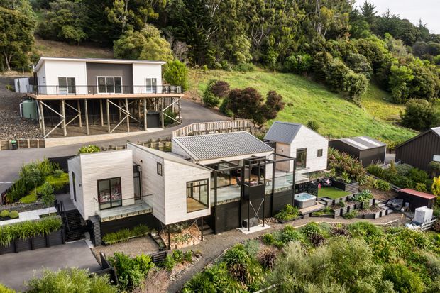 Aerial view of a contemporary multi-building residential property set into a densely vegetated, sloping site. The main low-rise home has light horizontal cladding, dark framed glazing, multiple pitched metal roof forms, terraces and landscaped garden areas. A separate elevated building and several detached outbuildings are visible, connected by paved paths and retaining structures.