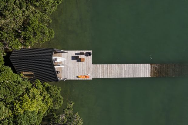 Aerial top-down view of a dark-roofed waterfront structure extending into calm green water, with a long timber deck or jetty furnished with outdoor seating, umbrellas, and a small orange kayak. Dense trees frame the shore-side end of the structure.